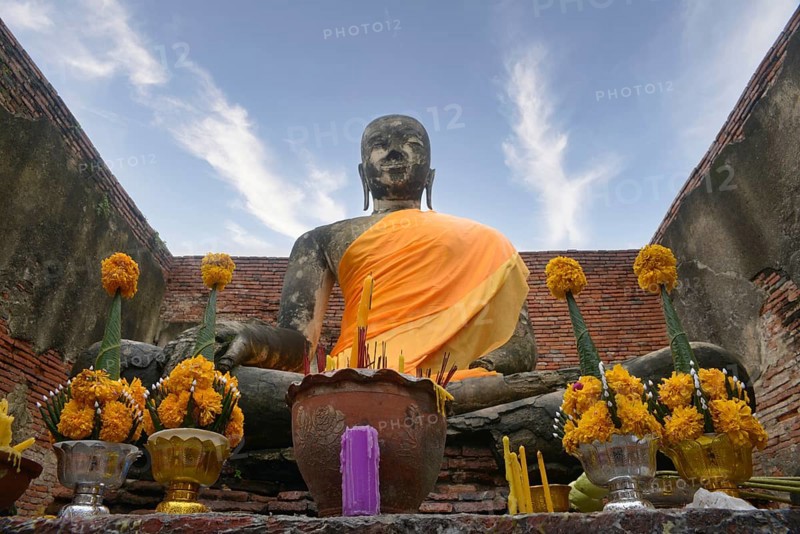 Majestic Buddha statue draped in orange cloth, surrounded by yellow flower offerings in Chiang Mai, Thailand. Set against a blue sky within ancient temple walls, it exudes peacefulness in Ayutthaya