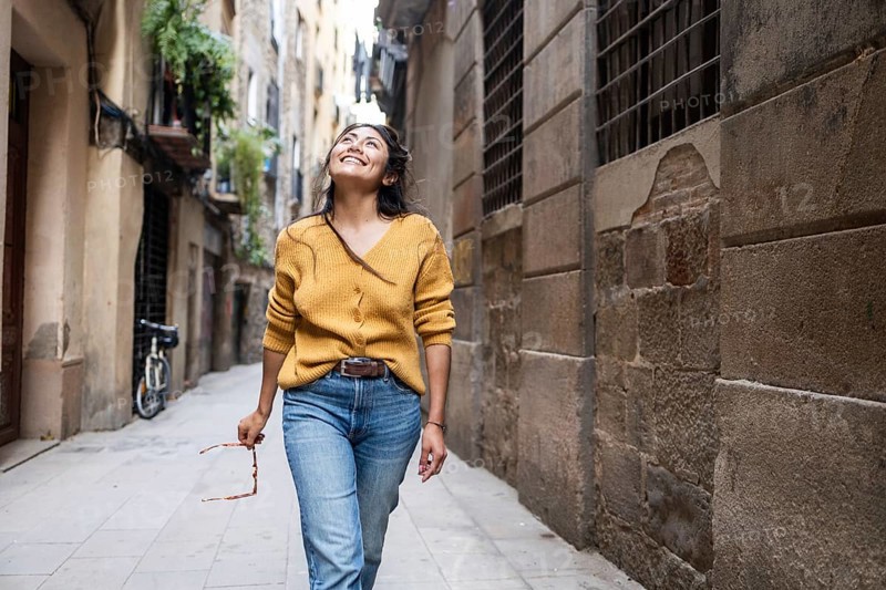 A Latin woman in a mustard sweater and jeans walks joyfully through a rustic urban alley, holding sunglasses. Her smile brightens the narrow, picturesque street