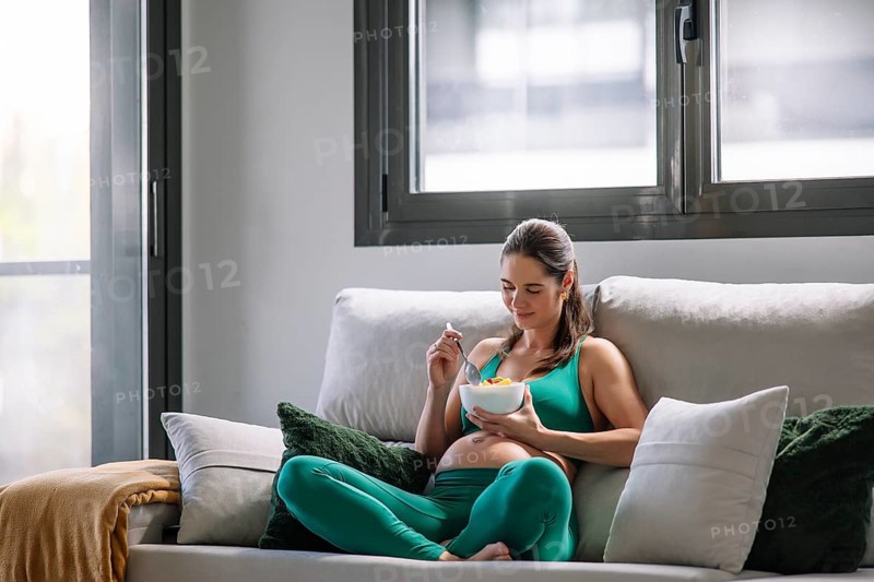 A pregnant woman sits comfortably on a couch in a yoga outfit, enjoying a relaxing moment while eating. Natural light from the window enhances the serene atmosphere of wellness