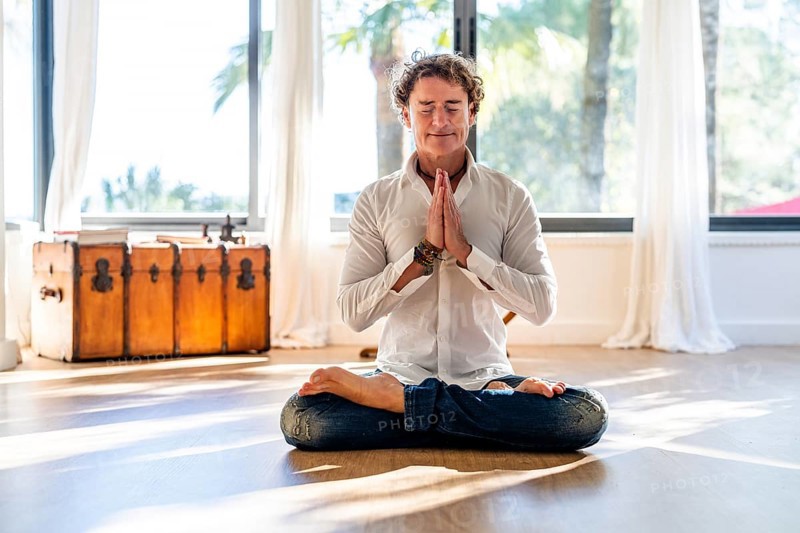 A calm man with curly hair meditates with eyes closed, seated in a lotus pose, hands in prayer position in a sunlit room decorated with minimalistic style and large windows