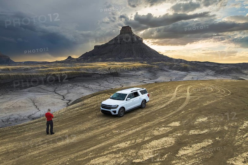 From above back view of unrecognizable man standing near a white SUV at Factory Butte, Utah, as the sun sets over the rugged landscape, showcasing dramatic skies and intricate ground patterns