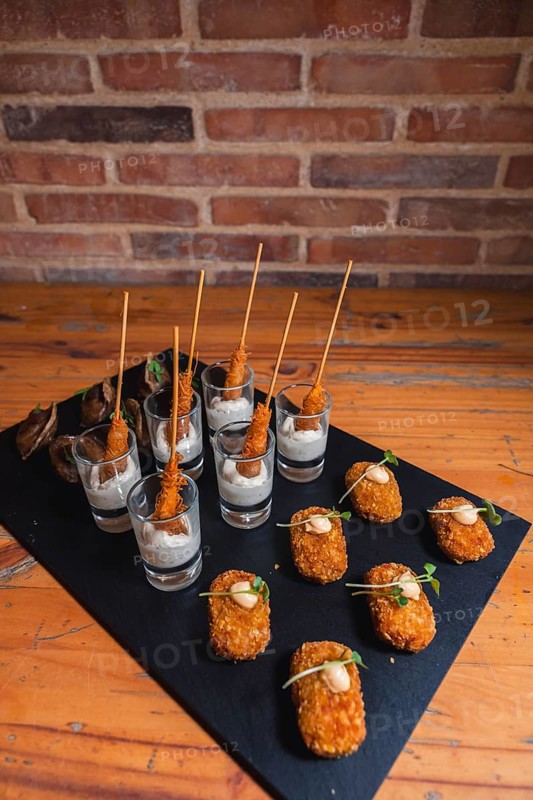 Elegant black slate platter featuring gourmet skewered appetizers in shot glasses with creamy dipping sauce, and crispy bite-sized snacks garnished with microgreens on a rustic table