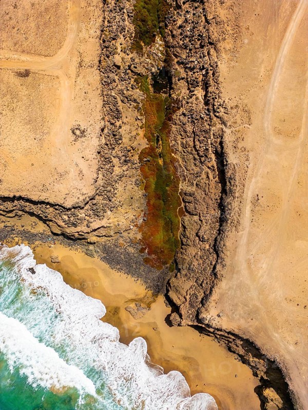 From above, an aerial shot captures a striking natural pool nestled within the rugged coastal landscape of Tindaya Beach in Fuerteventura, Canary Islands