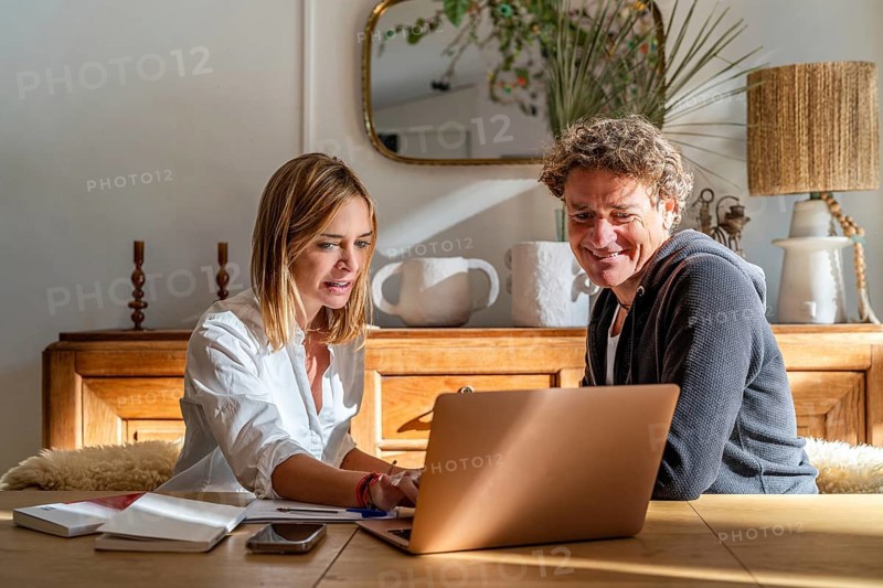 A couple works together at home, focused on a laptop The setting is cozy with a warm atmosphere, featuring a wooden table and modern decor Perfect for remote work themes