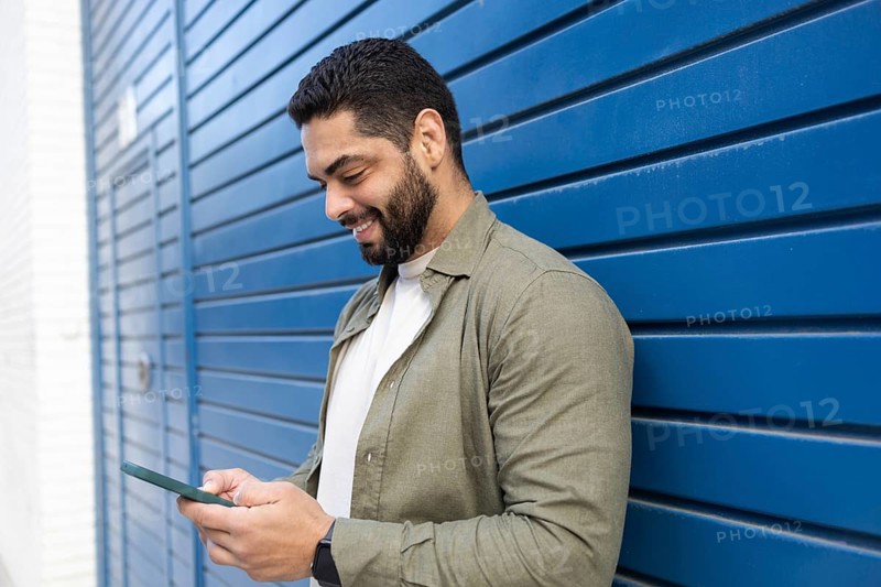 A man stands against a blue wall, smiling as he looks at his smartphone in the city. He wears a casual green shirt and a watch, emphasizing a relaxed and modern lifestyle