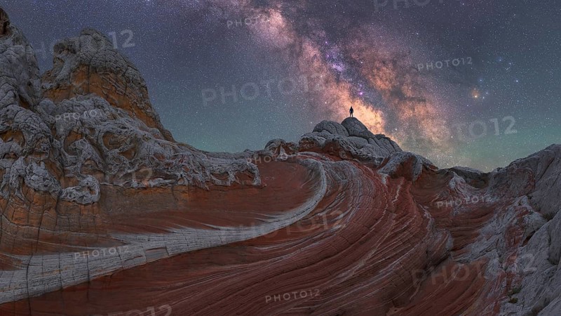 A stunning nightscape of the Milky Way galaxy arching over the unique rock formations at White Pocket in Arizona, US, with a man standing silhouetted against the stars
