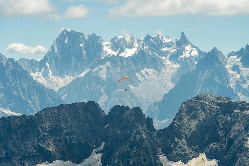 A lone paraglider glides gracefully above the rugged peaks of the French Alps near Chamonix and Mont Blanc, surrounded by dramatic snow-capped mountains under a clear blue sky