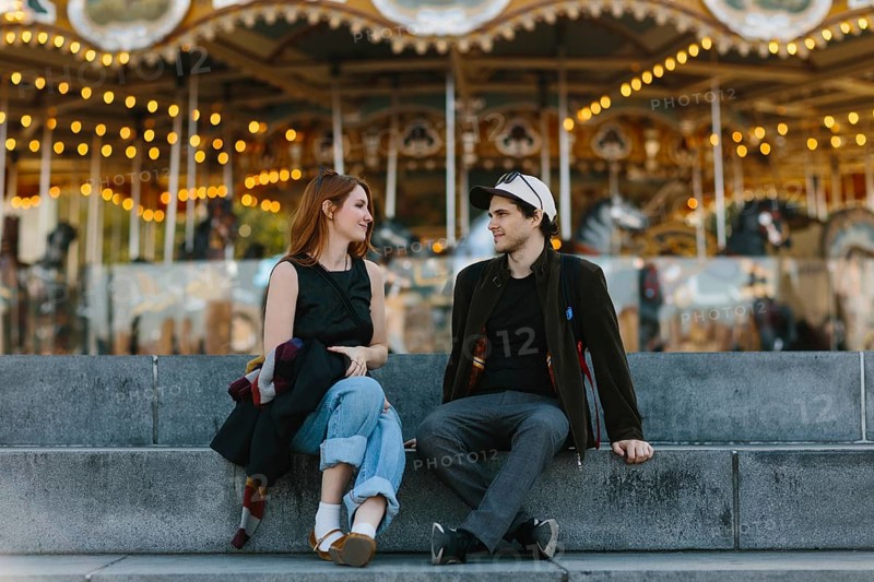 A young couple sits on stone steps, engaged in conversation, with a vibrant carousel in the background in Brooklyn Bridge Park. The warm lights create a cheerful, nostalgic atmosphere