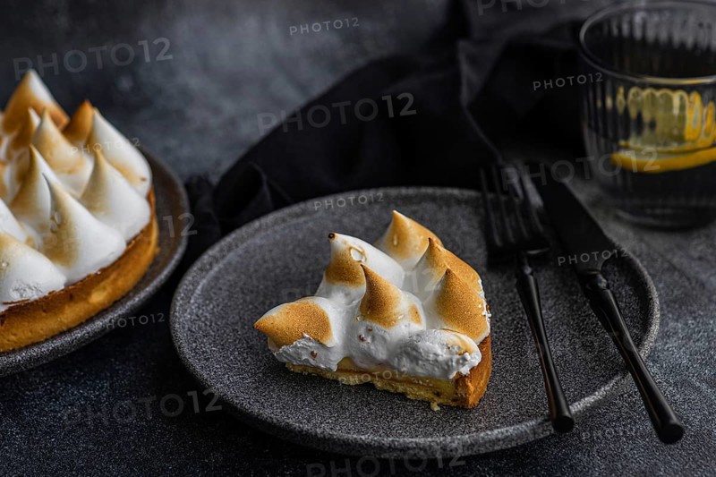 A slice of a homemade lemon cake sits elegantly on a dark plate, accompanied by a fork and knife. A glass with a slice of lemon adds a refreshing touch to the minimalist scene