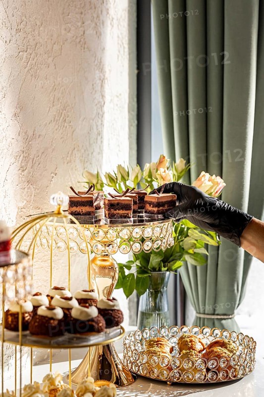A luxurious dessert spread featuring various cakes and pastries displayed on elegant, crystal adorned stands with a hand reaching for a chocolate treat. Lighting and flowers enhance the ambiance