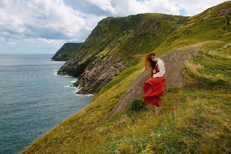 A young woman wearing a flowing red dress and white blouse explores the rugged, green hillside overlooking the ocean at Middle Cove, Newfoundland & Labrador, Canada. Her red hair is tousled by the wind