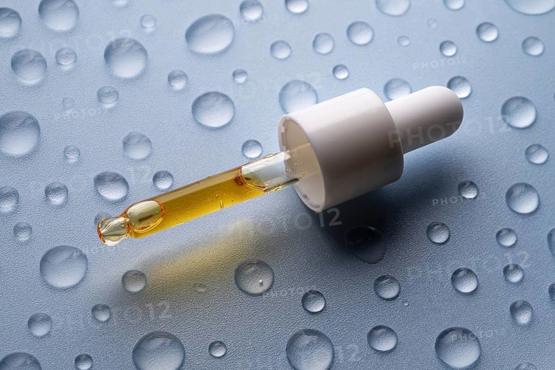 A close-up of a pipette bottle containing vitamin C serum, placed on a surface with scattered water droplets This image highlights the serum's hydrating properties