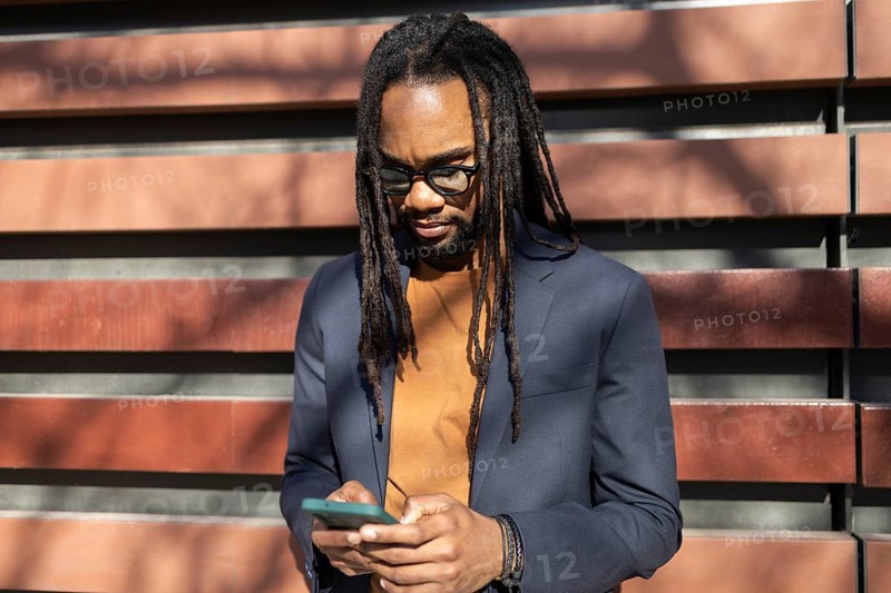 A cuban business professional with glasses and dreadlocks stands outdoors in sunlight, using a smartphone while wearing a smart blazer, against a modern wall backdrop