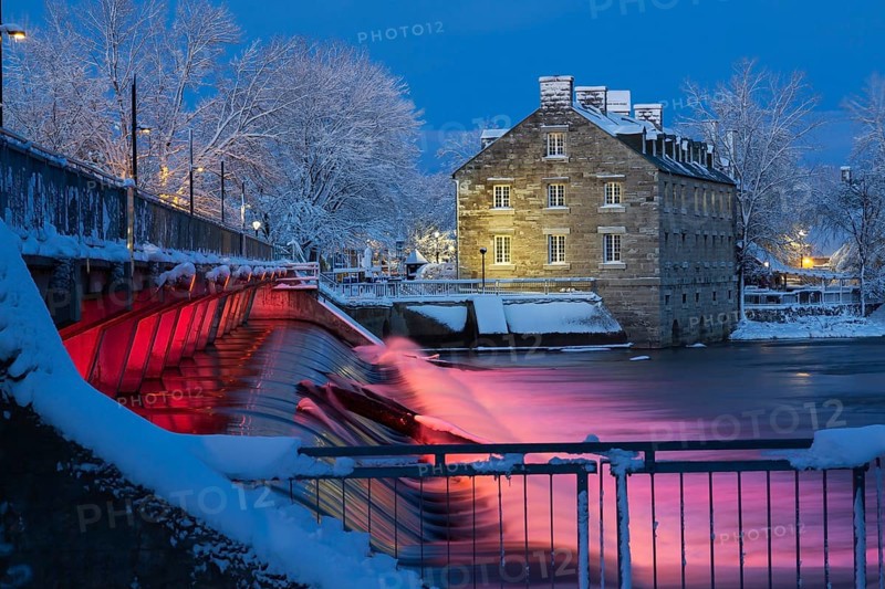 Illuminated Moulin Neuf water flow control dam and walkway over Des Mille-Iles river plus New Mill on Ile des Moulins with first winter like snow in late autumn at dusk, Old Terrebonne, Quebec, Canada