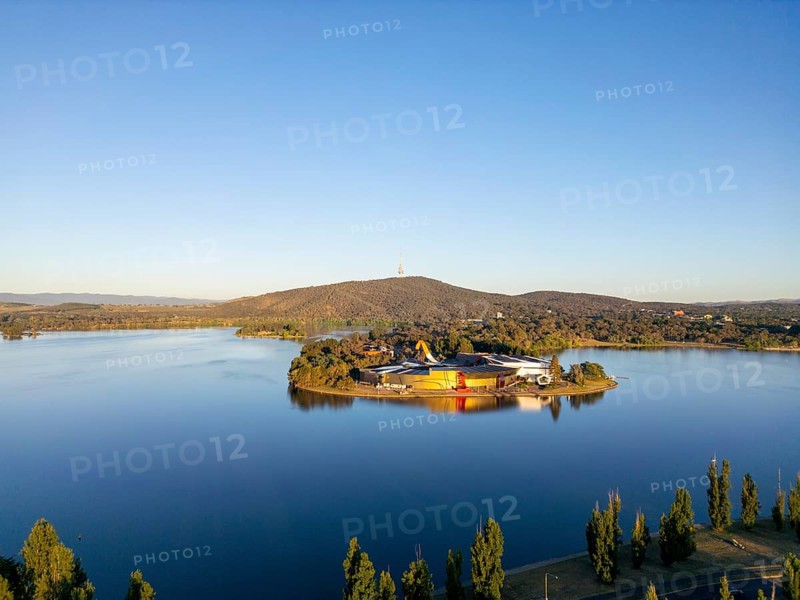 From above, this drone view of Canberra, the capital of Australia, featuring the serene waters of Lake Burley Griffin. The scene also includes a lush island hosting modern architectural structures and bordered by urban development