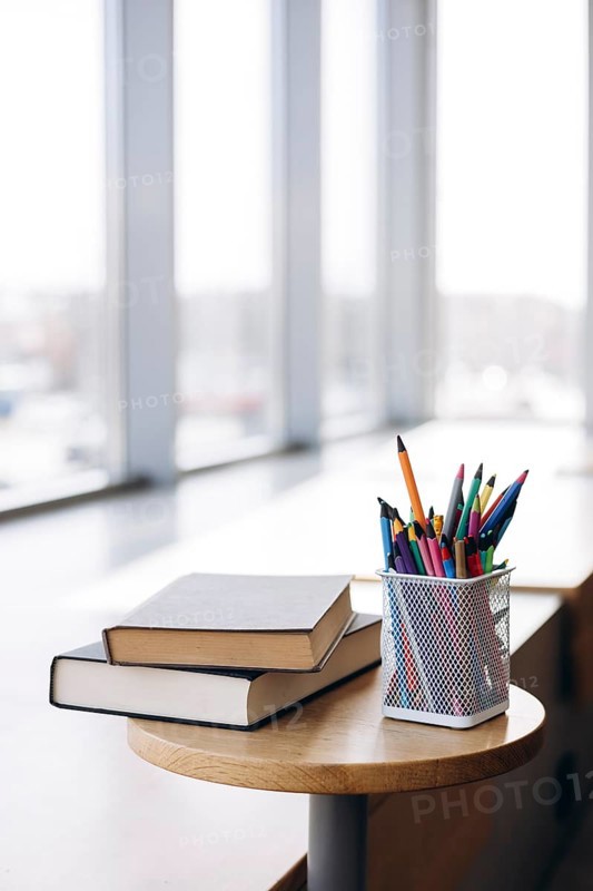 Books with dark covers stacked atop a circular wooden table beside a mesh pencil holder filled with colorful pencils, placed near a large window letting in natural light
