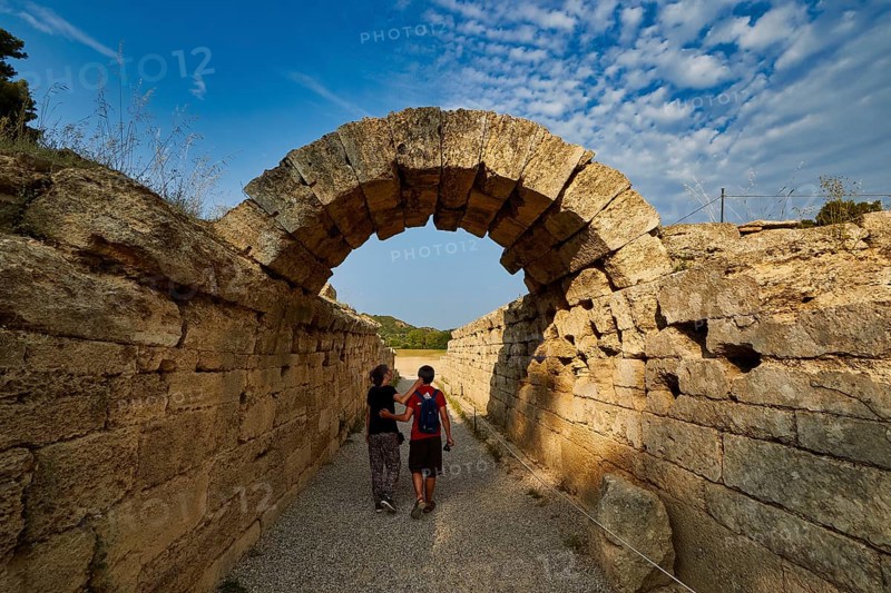 Crypt, vaulted passage to the stadium, two people walking through an ancient stone arch pass under a blue sky, Archaeological Site, Ancient Olympia, Olympia, Peloponnese, Greece