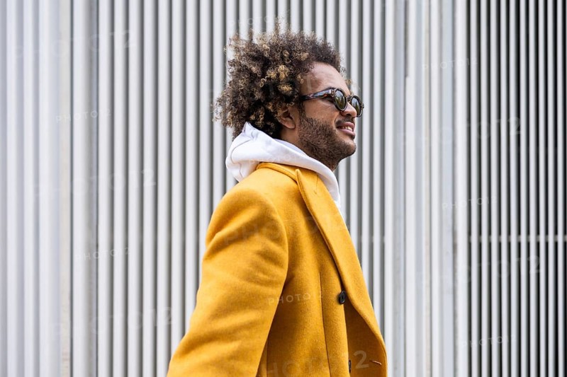 Afro man wearing a stylish mustard coat gazes confidently to the side. His sunglasses reflect a sense of cool sophistication against a backdrop of white vertical lines