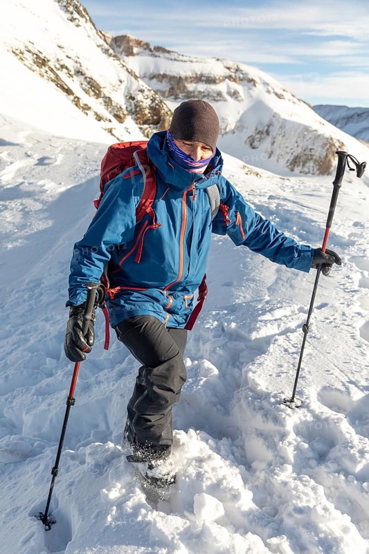 Adventurer in blue jacket and red backpack enjoys ski touring on a sunny day. Using poles for balance, trekking through fresh snow in a mountain landscape