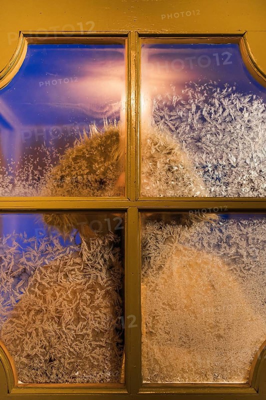 Frost-covered window with a blurred figure seen through the glass, capturing the essence of a cold winter day in Swedish Lapland. Warm light contrasts icy patterns