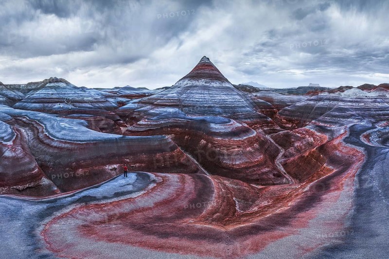 From above aerial drone photograph of an unrecognizable traveler standing on the vibrant and textured geological layers of Hanksville, Utah's landscape under a cloudy sky