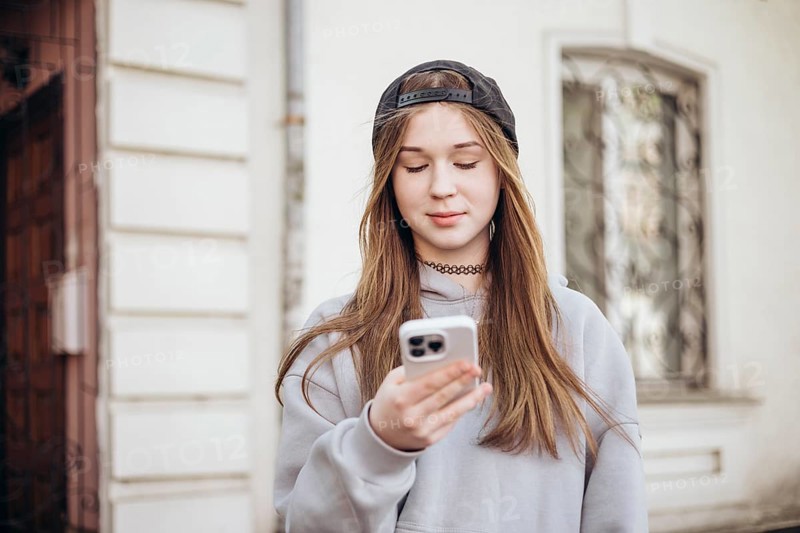A teenage girl wearing a hoodie and cap is focused on her smartphone while standing outside. She is engaged with her device, representing modern youth culture and technology use