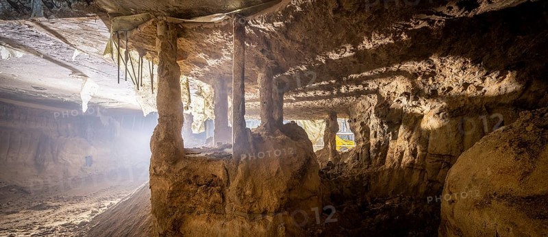A deep underground excavation reveals exposed soil columns and eroded concrete structures. The dim lighting and dust-filled atmosphere highlight the ongoing construction and reinforcement process
