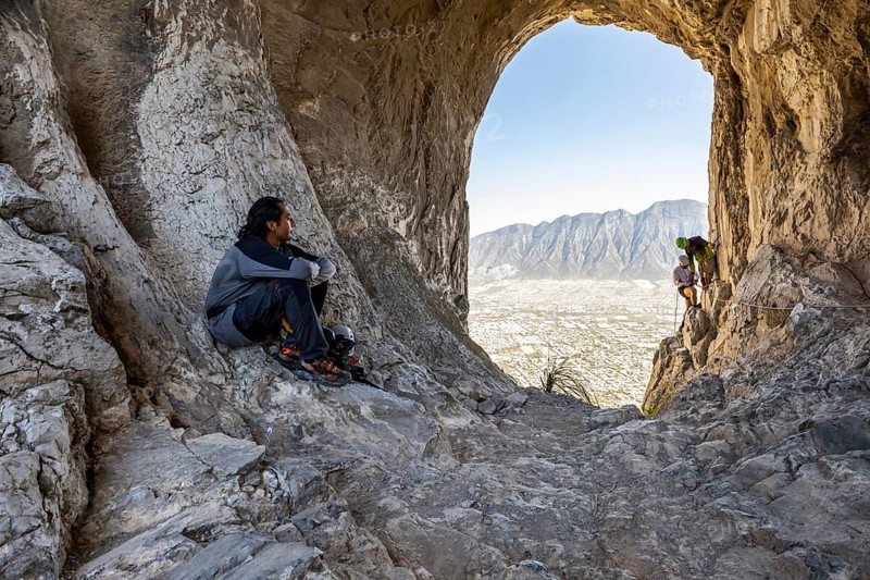 A climber rests on rocky terrain while two others practice mountaineering and rappelling in Eagleâ€™s Nest, Monterrey, Mexico. The cave opening frames the scenic mountains