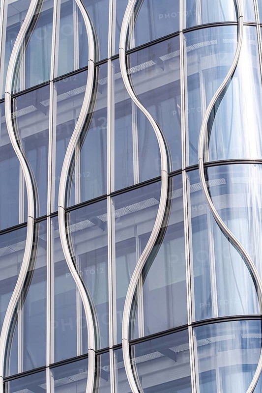 Close-up of a modern building facade, showcasing wavy glass panels and sleek metal frames. The undulating design creates a dynamic and futuristic architectural detail