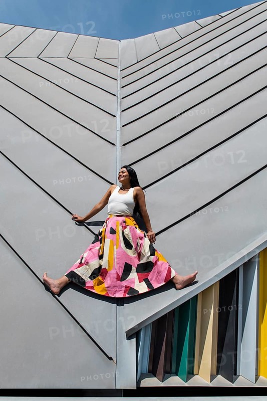 A woman wearing a vibrant, colorful outfit poses against striking modern architecture, showcasing a captivating contrast of urban design and fashion creativity