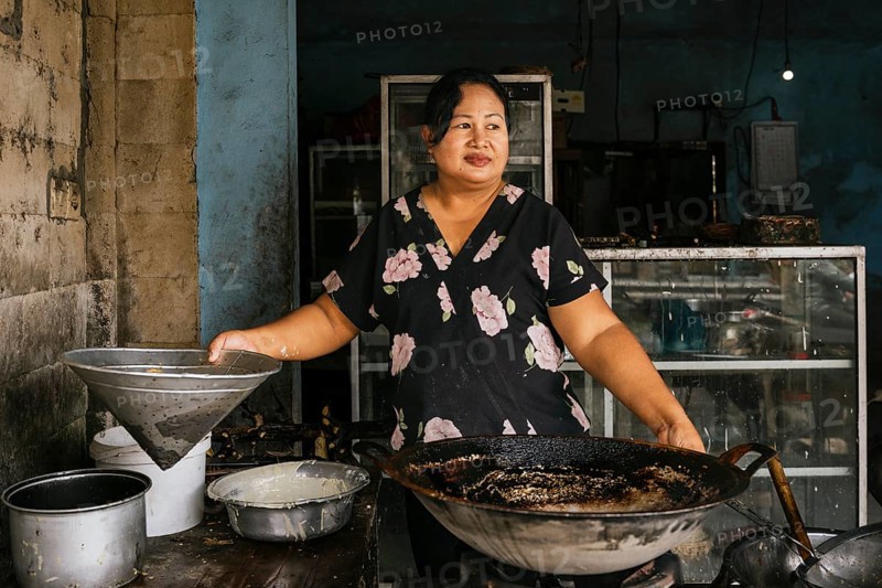 A woman stands in a rustic kitchen, preparing traditional dishes with a large wok. The worn walls and rustic tools create an authentic atmosphere, showcasing local cuisine of Bali