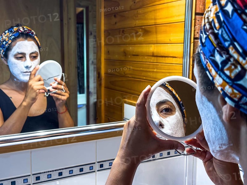 A woman examines her skincare routine using a hand mirror in a cozy bathroom of Victoria, Australia. She wears a white face mask and colorful headscarf, emphasizing personal care and relaxation
