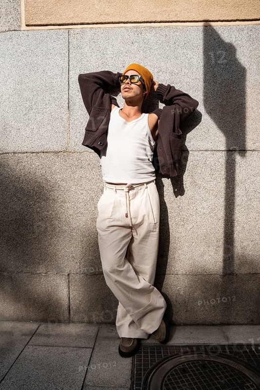 Stylish Colombian gay man wearing sunglasses and a beanie, leans against a city wall. His casual yet trendy outfit includes a tank top, cardigan, and wide-leg trousers