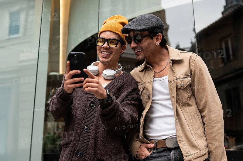 A joyful gay couple shares a moment of laughter while looking at a smartphone. They stand together outside, dressed casually, radiating happiness and companionship