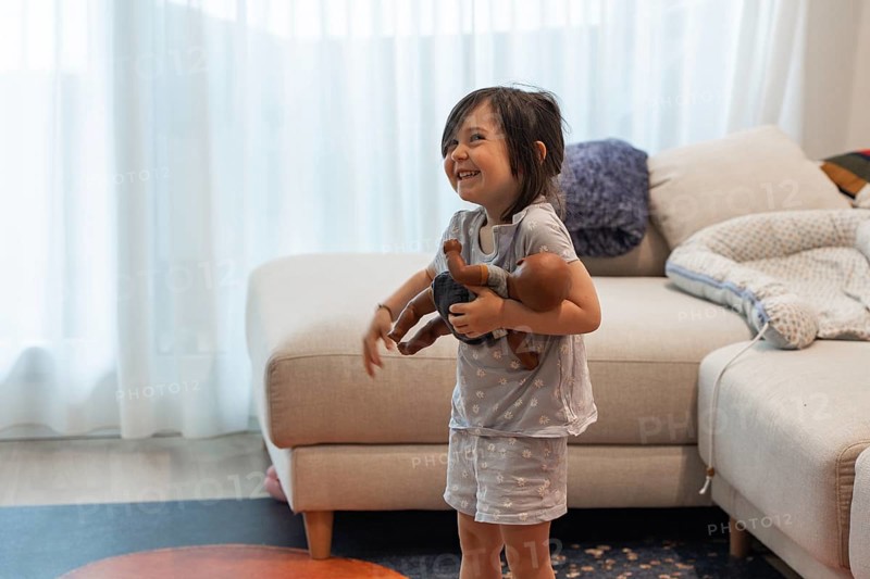 A young girl joyfully embraces her doll in a cozy living room, symbolizing love and innocence. The child's playful expression adds warmth to the vibrant home setting