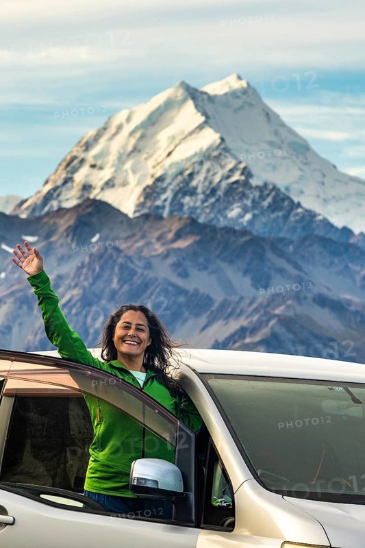 A woman stands joyfully in front of her car, surrounded by the breathtaking autumnal beauty of Mount Cook, New Zealand. The majestic mountain and vibrant colors enhance the scene