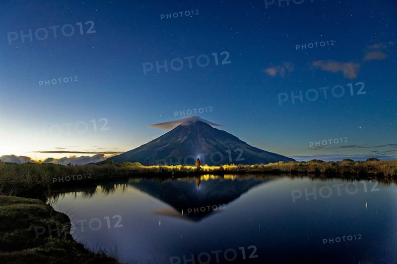 Back view of unrecognizable person gazing at Mount Taranaki's majestic peak reflected in a tranquil lake under a dawn sky, capturing New Zealand's serene natural beauty