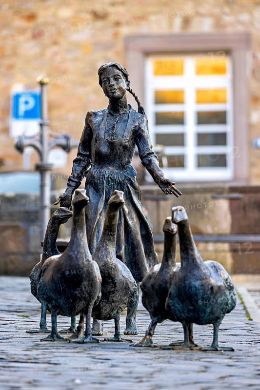 Bronze statue of a girl with geese on cobblestones, in the background a building with white windows, the Gänseliesel monument in Melsungen