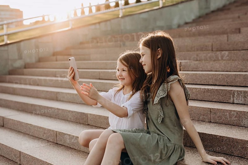 Two young girls sit on sunlit steps in summer, happily taking a selfie. They are enjoying a sunny day in a park, capturing moments of joy and togetherness with a smartphone