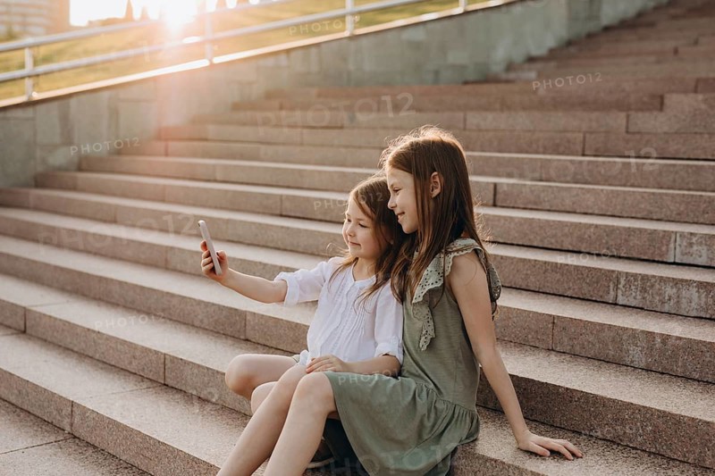 Two young girls sit together on sunlit stone stairs, smiling as they take a selfie. The warm glow of the sun highlights the joyful moment shared between friends in the city