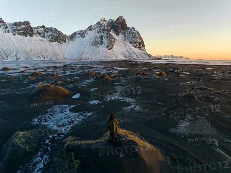 A man stands on a textured, frost-covered dune, gazing at the towering, snow-draped mountains of Stokksnes, Iceland, under a serene sunset sky