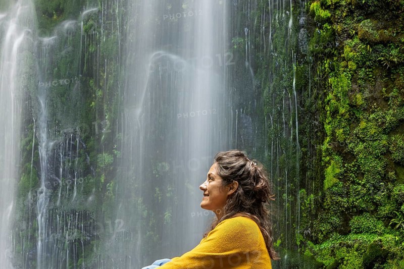 A woman in a yellow sweater enjoys a tranquil moment beside a lush green waterfall, surrounded by moss covered rocks. The scene evokes relaxation and nature beauty in the Great Ocean Road, Australia