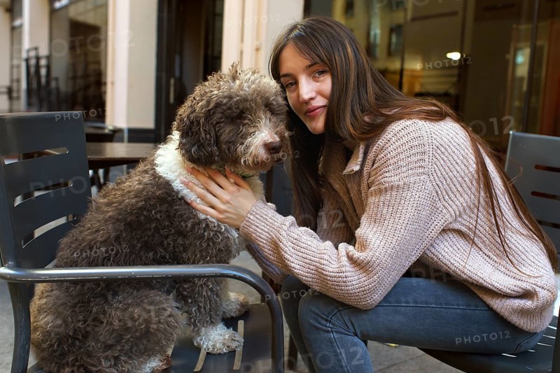 A young woman with long hair and a cozy sweater lovingly embraces her fluffy brown and white dog while sitting outdoors in a relaxed urban setting