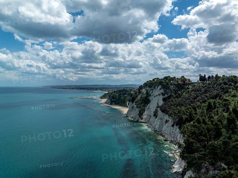 Aerial view of the stunning coastline in Sirolo, Italy. Dramatic cliffs rise from the turquoise sea, with lush greenery under a partly cloudy sky