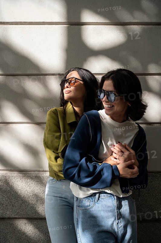 Two women, part of a lesbian couple, laughing and hugging in bright sunlight. Wearing casual outfits, they exude joy and warmth while enjoying a day outside