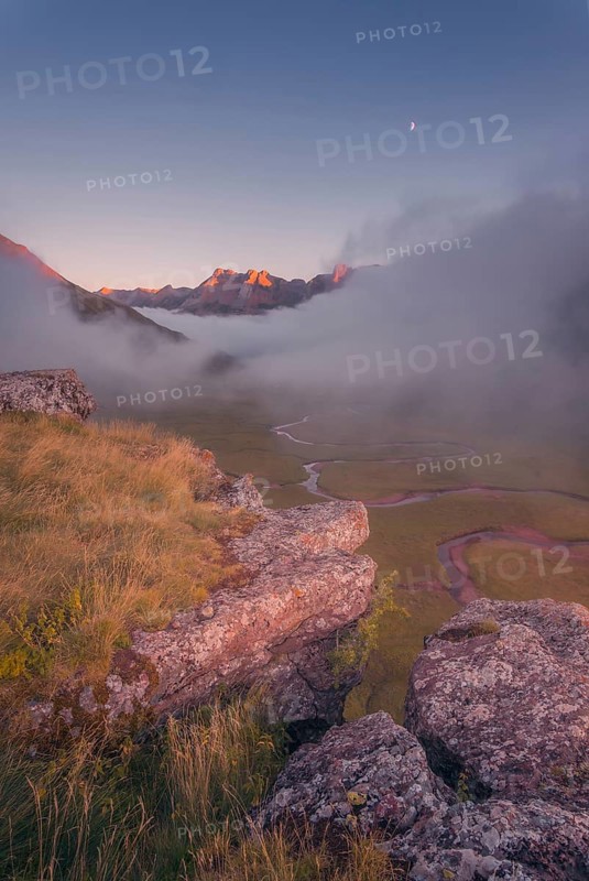 A breathtaking mountain landscape at dawn, with a rocky foreground, misty valley, winding stream, and glowing peaks in the Pyrenees. The moon hangs in the clear sky, adding tranquility