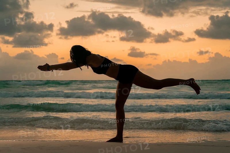 A woman balances gracefully in a yoga pose on a beach, silhouetted against a stunning sunrise. The scene captures tranquility, focus, and the beauty of nature in Tulum, Mexico