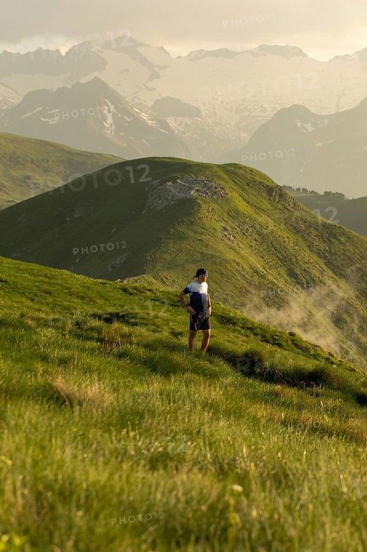 A man pauses to rest on a stunning mountain trail, surrounded by lush green hills under a serene sky, highlighting the harmony of outdoor adventure and nature
