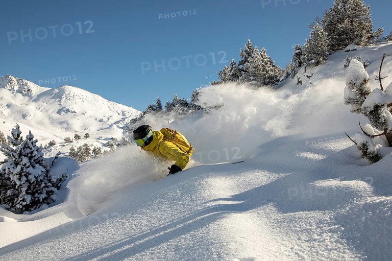 Dynamic freeride skiing in fresh powder snow with a skier in a yellow jacket navigating through a beautiful mountain landscape under a clear blue sky, capturing the thrill and excitement