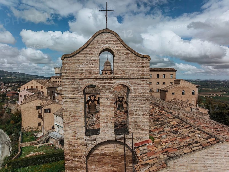 A historic bell tower stands amid rustic rooftops in Treia, Italy. The brick architecture contrasts with the lush countryside and blue skies in the background, ????, ????????, ??????